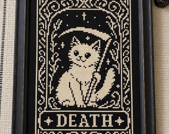 May include: A framed cross-stitch embroidery featuring a white cat holding a scythe, set against a black background with decorative borders and the word DEATH centered at the bottom in a rectangular frame, all enclosed in a black frame.