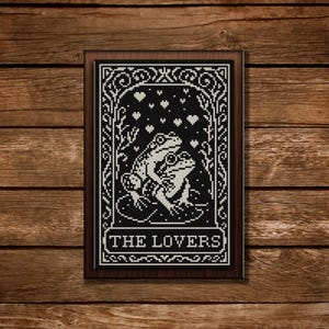May include: A framed cross-stitch depicting two white frogs on a lily pad, set against a black backdrop. The design includes hearts and floral motifs, with the text "THE LOVERS" at the base. The frame is dark brown, presented on a wooden surface.