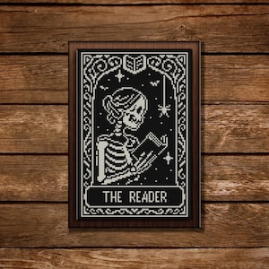 Puede incluir: Arte de punto de cruz en blanco y negro de un esqueleto leyendo un libro, enmarcado en un marco de madera oscura. El texto "THE READER" está en la parte inferior. El fondo es negro con estrellas y una araña.