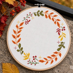 May include: A circular wooden embroidery hoop containing white Aida fabric with a cross-stitch pattern of an autumn wreath. The wreath features various leaves in shades of orange, yellow, and olive green, interspersed with small red berries.