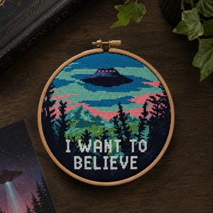 Könnte beinhalten: Ein runder Holzstickrahmen mit einem Kreuzstichmotiv, das ein dunkles UFO über einer Waldlandschaft mit rosa und blauen Wolken zeigt. Der Text I WANT TO BELIEVE ist in weißen Großbuchstaben am unteren Rand des Stickrahmens eingestickt.