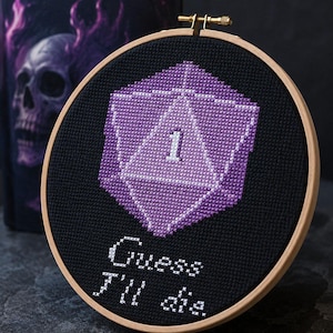 May include: A circular wooden embroidery hoop containing black fabric with a cross-stitched purple twenty-sided die featuring the number one in white. Below the die, the text "Guess I'll die" is stitched in white thread on the black fabric.