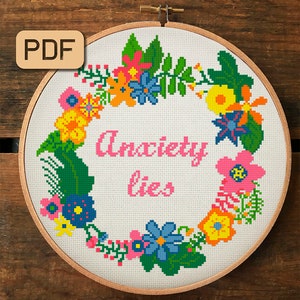 Może przedstawiać: Białe tamborek do haftu z kwiatowym wzorem wianka w jasnych kolorach, w tym różowym, żółtym, zielonym i niebieskim. Tekst "Anxiety lies" jest wyszyty na różowo w środku tamborka.