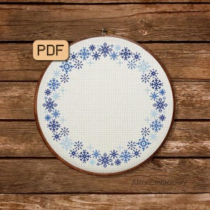 Könnte beinhalten: Ein weißer Stickrahmen mit einem blauen und weißen Schneeflockenrand. Der Rahmen befindet sich auf einem braunen Holzgrund. Der Text "PDF" befindet sich in einem braunen Rechteck auf der linken Seite des Bildes.