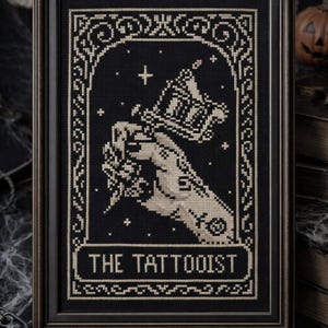 Peut inclure: Une broderie au point de croix encadrée sur tissu noir représentant une main tenant une machine à tatouer en fil crème. L'image est entourée d'une bordure ornée avec des petites étoiles et le texte THE TATTOOIST inscrit en bas en lettres capitales.