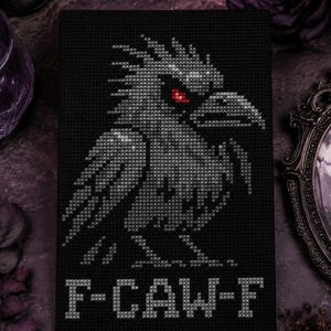 May include: A rectangular black cross-stitch embroidery on fabric featuring a grey bird with a sharp beak and a single glowing red eye. Below the bird, the text F-CAW-F is stitched in grey thread. The design is composed of visible X-shaped stitches.