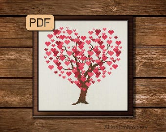 May include: Cross-stitch art of a tree with a heart-shaped canopy filled with pink and red hearts. The tree has a brown trunk and branches. The artwork is framed in a dark brown frame and set against a wooden background. The top left corner has a PDF icon.