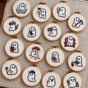 May include: A collection of fifteen circular wooden embroidery hoops, each containing a unique pixel art cross-stitch design of a white ghost engaged in various activities, including holding items like a rainbow flag, a football, or a wine glass.