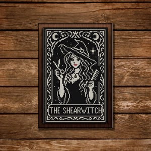 May include: Black and white cross-stitch art of a witch holding scissors and a comb, wearing a witch hat. The text "THE SHEARWITCH" is at the bottom of the framed artwork. The artwork is displayed on a wooden background.