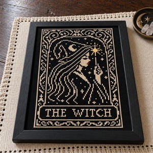 May include: A framed cross-stitch embroidery featuring a profile of a figure wearing a pointed hat, holding a star-tipped wand. The design is rendered in cream thread on a black background, with the text THE WITCH centered at the bottom.