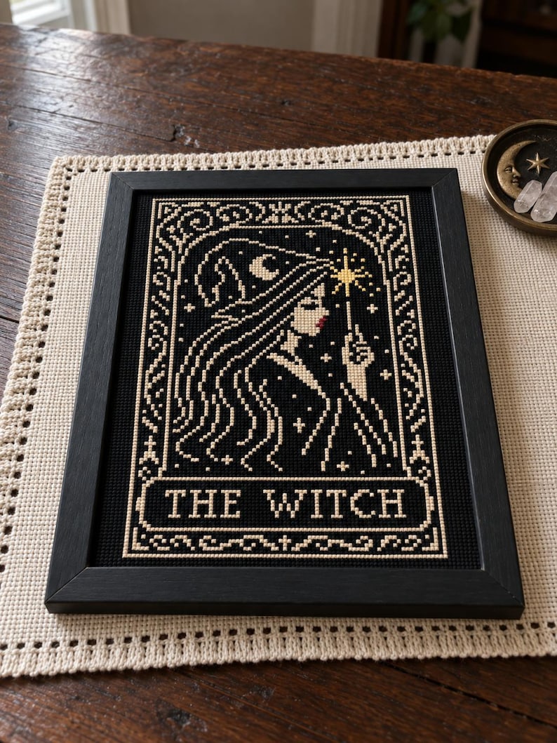 May include: A framed cross-stitch embroidery featuring a profile of a figure wearing a pointed hat, holding a star-tipped wand. The design is rendered in cream thread on a black background, with the text THE WITCH centered at the bottom.