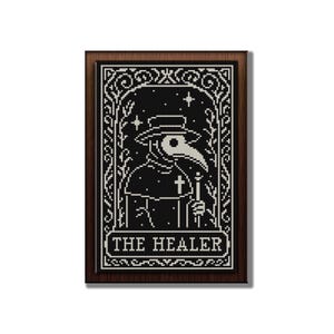 May include: A framed black and white cross-stitch design featuring a plague doctor with a long beak, hat, and cross. The text "THE HEALER" is at the bottom. The frame is a dark brown color.