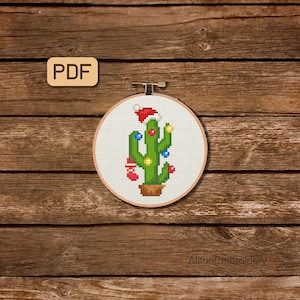 May include: A cross-stitch pattern of a cactus wearing a Santa hat and decorated with Christmas lights and ornaments. The pattern is in a white embroidery hoop on a wooden background.