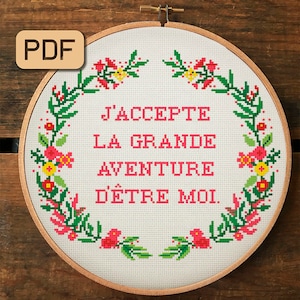 May include: A white embroidery hoop with a floral wreath design and the French phrase "J'accepte la grande aventure d'être moi." in pink lettering.