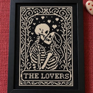 May include: A framed cross-stitch embroidery featuring two skeletons embracing within an ornate border. The design is worked in cream thread on a black fabric background. The text THE LOVERS is stitched in a rectangular box at the bottom.