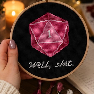 Op de afbeelding: Kruissteekborduurwerk in een houten ring met een roze en wit geometrisch ontwerp met het nummer 1 op een zwarte achtergrond. De tekst "Well, shit." is eronder geborduurd. De ring wordt door een persoon vastgehouden.
