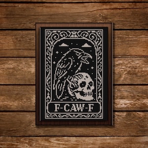 May include: Black and white cross-stitch art in a dark wooden frame. The design features a raven perched on a skull, with the text "F-CAW-F" at the bottom. The background is black with a decorative border.