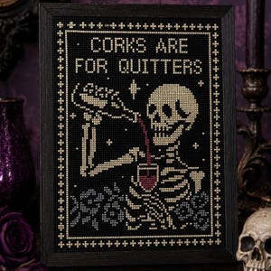 May include: A framed cross-stitch embroidery on black fabric featuring a skeleton pouring red wine into a glass. The text reads CORKS ARE FOR QUITTERS in white capital letters. The design includes small white stars and a decorative border.