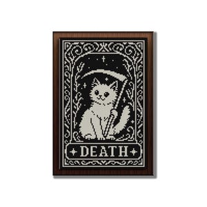 May include: A framed cross-stitch embroidery featuring a white cat holding a scythe, set against a black background with white stars and floral borders. The word DEATH is printed in white capital letters within a rectangular banner at the bottom.
