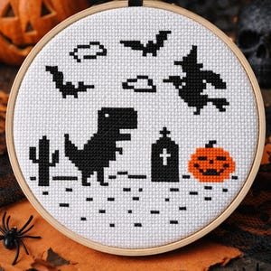 Può includere: Un telaio da ricamo circolare in legno contenente un pezzo di tessuto bianco con un disegno a punto croce. Il motivo presenta un dinosauro nero pixelato, un cactus, una lapide, due pipistrelli, una strega su una scopa e una zucca arancione.
