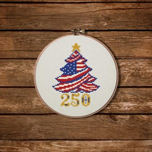 Pode incluir: Bordado em ponto cruz em um bastidor redondo. O design apresenta uma árvore de Natal nas cores da bandeira americana, com uma estrela dourada no topo. O número "250" é bordado em ouro abaixo. O fundo é uma superfície de madeira.