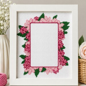 May include: A white framed cross-stitch featuring a floral design. The embroidery showcases pink and magenta flowers with green leaves, arranged around a central rectangular space. The frame is white, and the overall aesthetic is delicate and handcrafted.