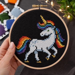May include: A cross-stitch embroidery of a white unicorn with a rainbow mane and tail, set against a black background. The unicorn is framed in a wooden hoop. Colorful embroidery floss is visible in the background.