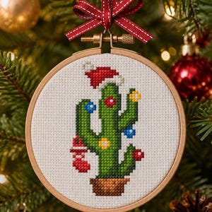 May include: A cross-stitch Christmas ornament featuring a green cactus wearing a Santa hat and decorated with colorful lights. The cactus is in a brown pot, set against a white background within a wooden embroidery hoop, and is suspended by a red ribbon.