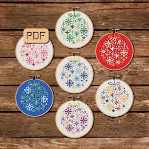 May include: Six colorful Christmas ornament embroidery patterns with a variety of snowflake designs. The patterns are displayed in embroidery hoops on a wooden background. The text "PDF" is visible in the upper left corner of the image.