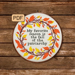May include: A cross-stitch embroidery hoop with a white background and a wreath of orange and yellow leaves. The text inside the wreath reads "My favourite season is the fall of the patriarchy".