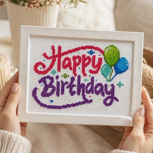 May include: A framed cross-stitch embroidery featuring the text Happy Birthday in pink and purple thread. The design includes three small balloons in green, light blue, and dark blue, along with tiny colourful star accents on a white fabric background.
