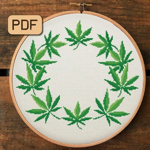 Puede incluir: Un aro de bordado blanco con un diseño de corona de hojas de marihuana verdes. El aro es de madera y tiene un acabado natural. El diseño es simple y moderno, y quedaría genial en cualquier hogar.