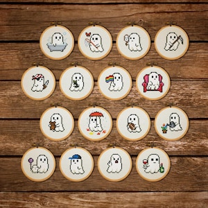 May include: A collection of 15 small, round embroidery hoops with cross-stitched ghost designs. Each ghost is depicted in a different scene, such as taking a bath, holding a balloon, or wearing a hat. The hoops are arranged on a wooden surface.