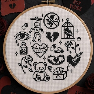 Pode incluir: Um bastidor de bordado circular de madeira contendo tecido branco com desenhos em ponto cruz preto, incluindo um esqueleto, um coração partido, um pássaro numa gaiola, um urso de peluche, comprimidos, uma rosa e uma garrafa marcada com um X.