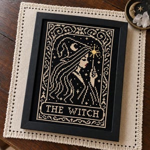 May include: A framed cross-stitch embroidery featuring a profile of a figure wearing a pointed hat, holding a star-tipped wand. The design is stitched in cream thread on a black background with the text THE WITCH centered at the bottom.