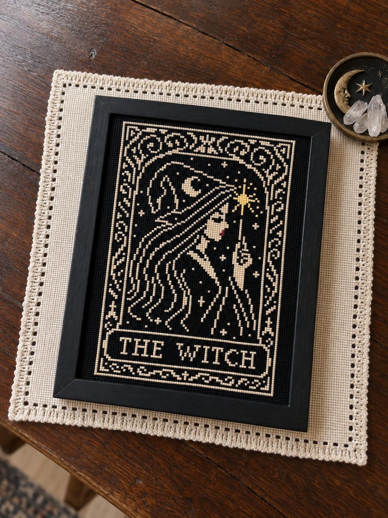 May include: A framed cross-stitch embroidery featuring a profile of a figure wearing a pointed hat, holding a star-tipped wand. The design is stitched in cream thread on a black background with the text THE WITCH centered at the bottom.