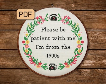 May include: A white embroidery hoop with a floral wreath design and the text "Please be patient with me I'm from the 1900s". The wreath is made of green leaves and pink and yellow flowers. There are two black rotary phones on either side of the text.