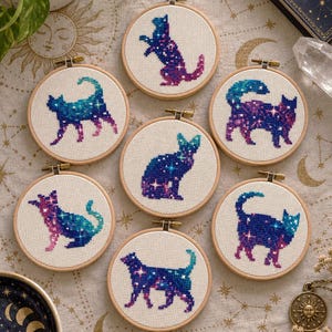 May include: Seven circular wooden embroidery hoops containing cross-stitch designs of cats. Each cat features a vibrant galaxy-inspired color gradient of blue, purple, and pink, accented with small white star-like stitches on white fabric.