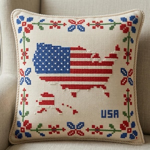Puede incluir: Cojín cuadrado con un diseño de punto de cruz que representa la bandera de EE. UU. y el contorno del mapa en rojo, blanco y azul. El cojín tiene un fondo color crema y un borde decorativo con detalles florales. La palabra "USA" está bordada en azul.