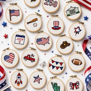 May include: A collection of seventeen circular wooden embroidery hoops featuring pixel art cross-stitch designs with American patriotic themes, including a bald eagle, the number 250, the White House, a flag, baseball gear, a top hat, and a rocket.