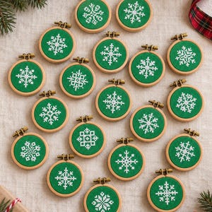 May include: A collection of small, round wooden embroidery hoops, each containing green fabric with a unique white cross-stitched snowflake pattern. Each hoop features a small metal tightening screw at the top, arranged in a scattered display.