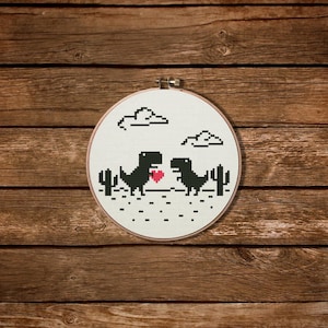 May include: A cross-stitch hoop art piece featuring two black pixelated dinosaurs facing each other, with a red heart between them. The design includes cacti, clouds, and a dotted ground, all on a white background. The hoop is set against a wooden backdrop.