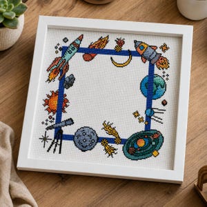 May include: A framed cross-stitch featuring a space-themed design. The artwork includes rockets, planets, stars, and a telescope, all set against a white background with a blue border. The frame is white, and the piece is displayed on a wooden surface.