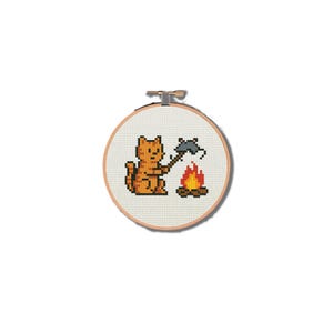 Könnte beinhalten: Ein Kreuzstich-Stickrahmen mit einer pixeligen orangefarbenen Katze, die eine Axt hält, mit einem Lagerfeuer. Die Katze befindet sich neben einem Lagerfeuer mit orangefarbenen und gelben Flammen. Das Design befindet sich auf einem cremefarbenen Stoff.