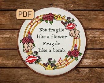Könnte beinhalten: Ein weißer Stickrahmen mit einem goldenen und roten Blumendesign. Der Rahmen zeigt den Text "Not fragile like a flower. Fragile like a bomb."