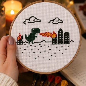 May include: A cross-stitch embroidery hoop featuring a pixelated design of a green dinosaur breathing fire onto a city skyline. The design includes black clouds and buildings, with red and orange flames. The hoop is held by a person, with colorful embroidery floss and scissors in the foreground.