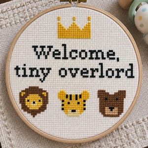 May include: A circular wooden embroidery hoop containing white fabric with a cross-stitch design. The design features a yellow crown above the black text "Welcome, tiny overlord" and three animal faces: a lion, a tiger, and a brown bear.