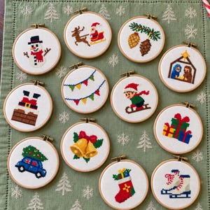 May include: A collection of twelve circular wooden embroidery hoops featuring pixel art cross-stitch designs of Christmas themes including a snowman, Santa in a sleigh, pinecones, a nativity scene, bunting, a sledder, Santa in a chimney, gifts, a car with a tree, bells, a stocking, and ice skates.