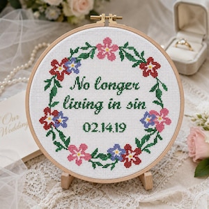Könnte beinhalten: Runde Kreuzstich-Stickerei in einem Holzreifen mit dem Text "No longer living in sin" und dem Datum 02.14.19. Das Design umfasst einen grünen Blumenkranz mit roten, rosa und blauen Blumen. Die Stickerei wird auf einem kleinen Holzständer präsentiert.