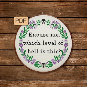 Puede incluir: Un aro de bordado blanco con un diseño floral verde y morado y el texto "Excuse me, which level of hell is this?" El aro está sobre un fondo de madera marrón.